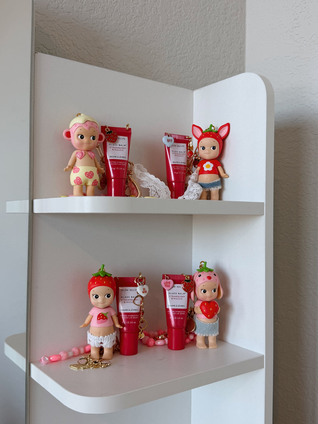Small dolls and containers on a white corner shelf against a neutral wall.